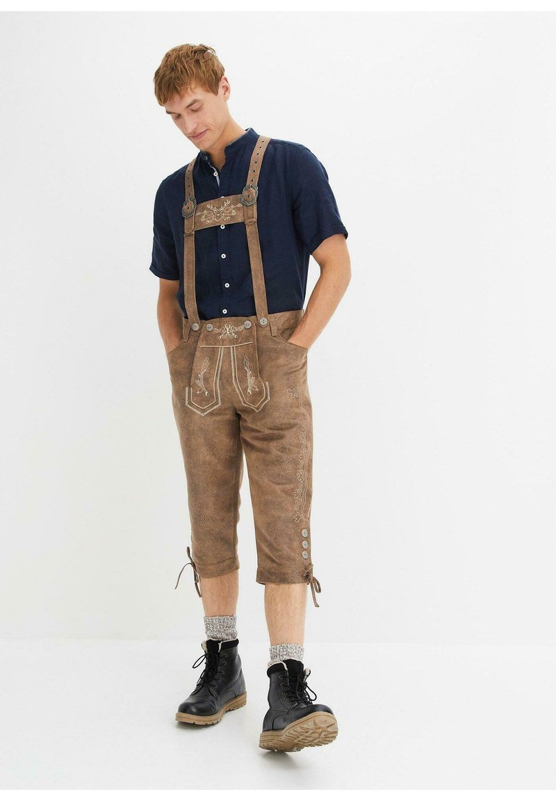 Lederhosen in pelle marrone con dettagli ricamati, spalline regolabili, due tasche e orli arrotolati. Abbinati a una camicia scura e stivali neri.