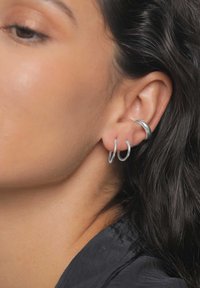 Sølv øreringe i hoopdesign i forskellige størrelser stablet i øret, med glatte overflader og et minimalistisk design. Håret er bølget og mørkt.