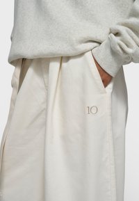 Sweat-shirt gris clair avec poignets côtelés, associé à un pantalon large couleur crème avec un discret "10" brodé près de la poche.