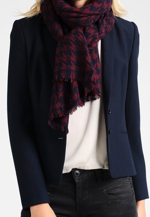 Femme portant un blazer bleu foncé, un haut blanc, un jean noir et une épaisse écharpe à motif pied-de-poule rouge et marine enroulée autour du cou.