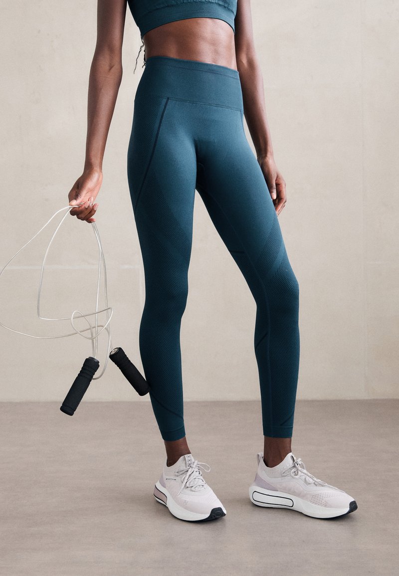 Teal hoge taille legging met gestructureerde patronen, gecombineerd met lichtgrijze sportschoenen. Houdt een springtouw vast met zwarte handvatten.