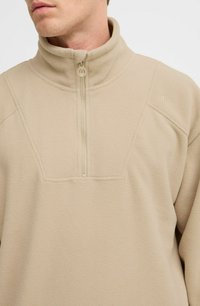 Beige fleece pullover med høj krave, halv lynlås og struktureret overflade, med en stikdetalje på forsiden og logo på ærmet.