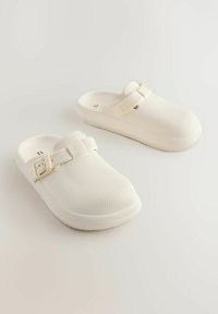 Witte klompen met textuur, een afgeronde teen, platte zool en verstelbare riem. Gemaakt van lichtgewicht materiaal, met een geribbeld ontwerp.