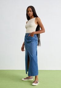 Top sin mangas beige, falda vaquera azul de talle alto con una abertura lateral y zapatos grises sin cordones. Materiales suaves y texturizados.