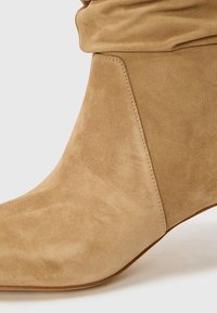 Stivaletto beige in suede con un design sovrapposto, punta affusolata e una texture liscia. Caratterizzato da una cucitura a vista per un tocco di dettaglio in più.