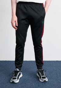 Svarta träningsbyxor med röda sidostripes, gjorda av slät tyg; elastisk midjeband. Bärs med svarta och grå sneakers.