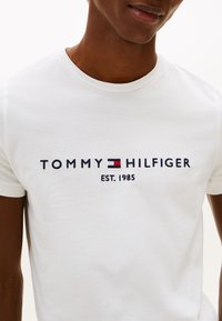 Valge puuvillane t-särk tumesinise tikitud tekstiga "TOMMY HILFIGER EST. 1985" ja väikese punase ning sinise logodetailiga. Klassikalise ümarkaelusega.