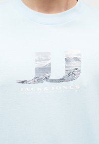 Ανοιχτό μπλε πουκάμισο με μεγάλο λογότυπο JJ που απεικονίζει ορεινό τοπίο και το κείμενο "JACK & JONES" κάτω από το στήθος.