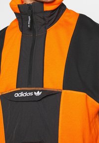 Giacca a pullover arancione e nera con collo alto, mezza cerniera frontale e logo Adidas ricamato sul petto. Presenta un design con pannelli a contrasto.