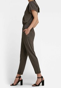 Mono caqui de manga corta, con cintura elástica, bolsillos laterales y puños enrollados. Combinado con sandalias negras de tacón cuadrado.