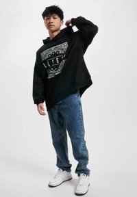 Black hoodie with a white graphic design, paired with blue denim jeans and white sneakers. The hoodie has a loose fit and a kangaroo pocket.