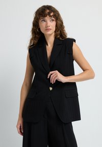 Černý bezrukávový blejzr s širokými klopami, texturovanou tkaninou, dvěma předními kapsami a zlatým knoflíkem ve tvaru srdce uprostřed.