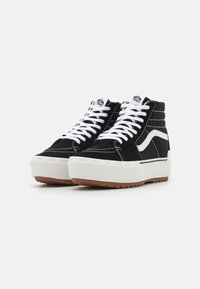 Czarne zamszowe sneakersy typu high-top z białymi sznurówkami, kontrastowym białym paskiem po boku, grubą teksturowaną gumową podeszwą i brązową podeszwą zewnętrzną.