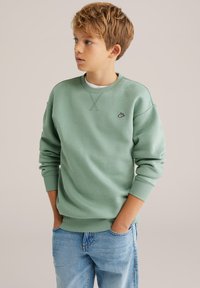 Mätovo zelená mikina s rebrovanými manžetami a lemom, s vyšívaným logom na hrudi. Kombinovaná so svetlomodrými džínsami.