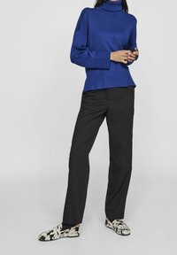 Maglione dolcevita blu con vestibilità comoda e polsini a coste, abbinato a pantaloni neri e scarpe slip-on con fantasia colorata.