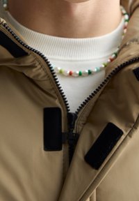 Camelkleurig jack met ritssluiting, zwarte klittenbandaccenten, gedragen over een witte coltrui. Kleurrijke kralenketting om de nek.