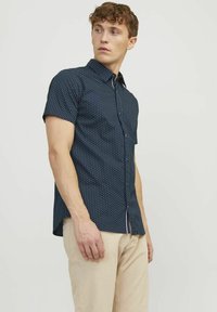Camicia a maniche corte di colore blu navy con motivo a pois bianchi, con bottoni davanti e orlo curvo. Indossata con pantaloni beige chiaro.