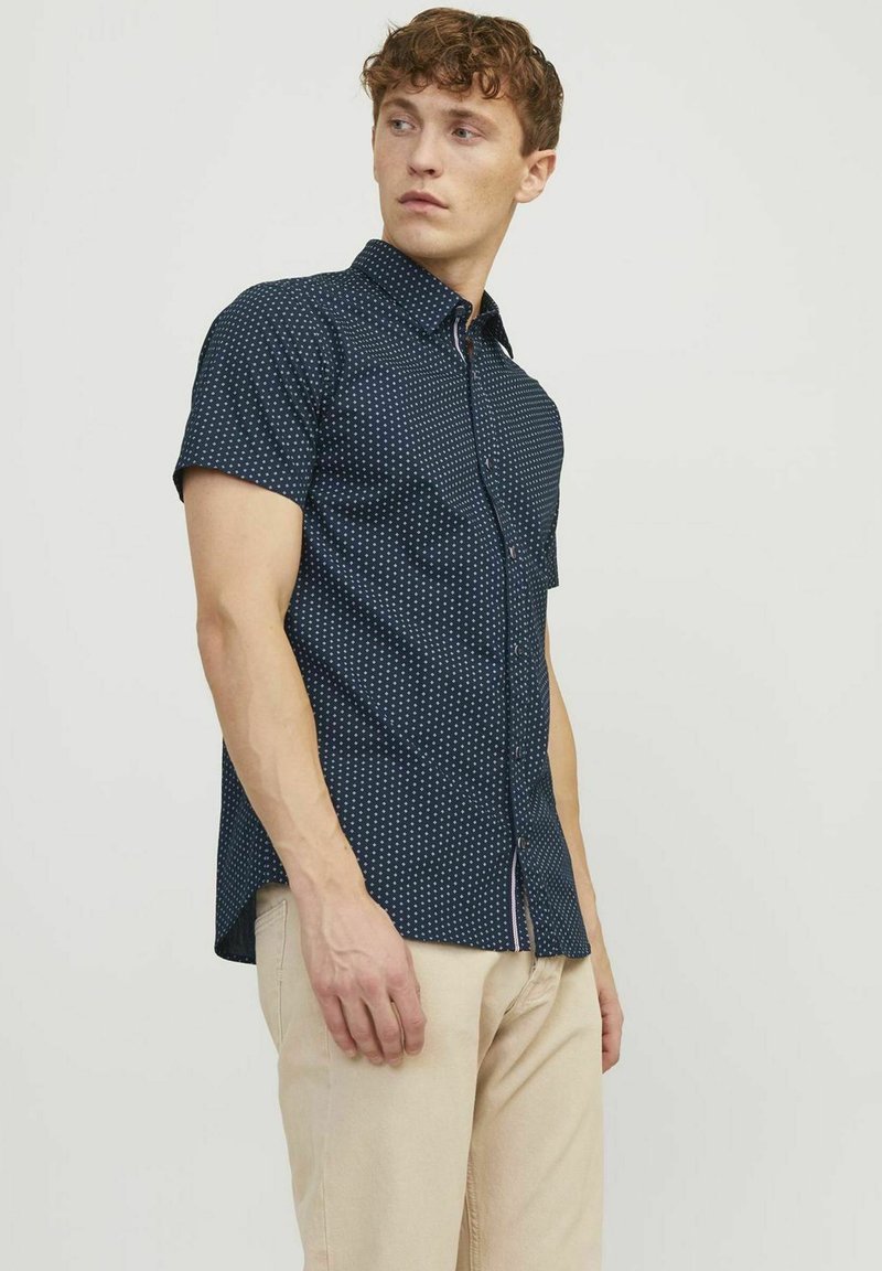 Camicia a maniche corte di colore blu navy con motivo a pois bianchi, con bottoni davanti e orlo curvo. Indossata con pantaloni beige chiaro.