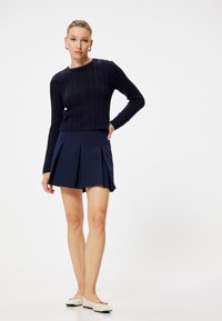 Pull marin avec un motif en maille torsadée, associé à une jupe plissée marine. Les chaussures sont beiges avec un accent décoratif. Design simple et épuré.
