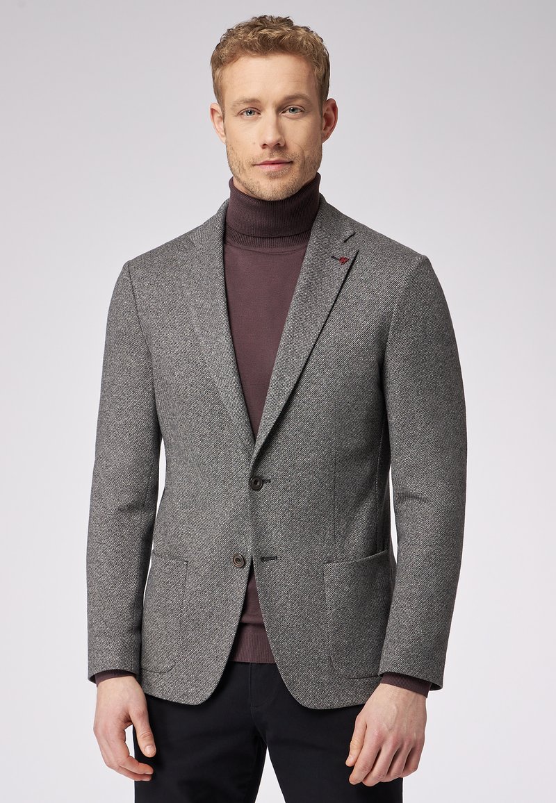 Gray houndstooth blazer made of textured fabric, featuring two front pockets, notch lapels, and two buttons. Worn over a dark turtleneck.