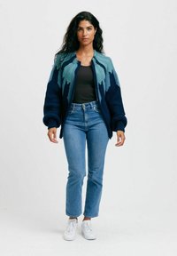 Stickad kofta i mörk marinblå med mönster av gröna och teal berg; texturerad tyg, avslappnad passform och ribbade ärmar. Kombination med ljusa jeans och vita skor.