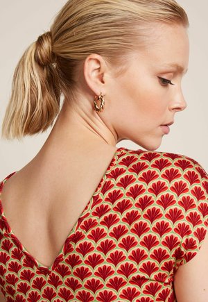 Blonde Frau mit zurückgebundenen Haaren, die goldene Creolen trägt und ein rotes Kleid mit Muster und tiefem V-Rücken, schaut seitlich nach unten.