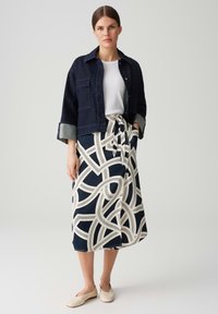 Veste en denim bleu foncé avec deux poches avant, associée à une jupe longue ornée de motifs abstraits crème sur fond marine. Chaussures légères.