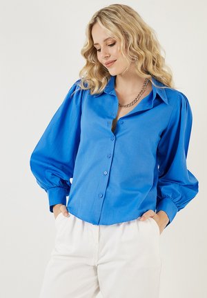 Blue button-up shirt with puffed sleeves and a collar. Smooth fabric with a slight sheen. Worn with light-colored trousers.