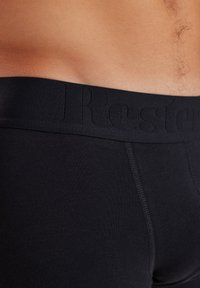 Boxer da uomo neri in cotone elastico con un elastico testurizzato nero che presenta un logo in rilievo. Superficie liscia e design aderente.