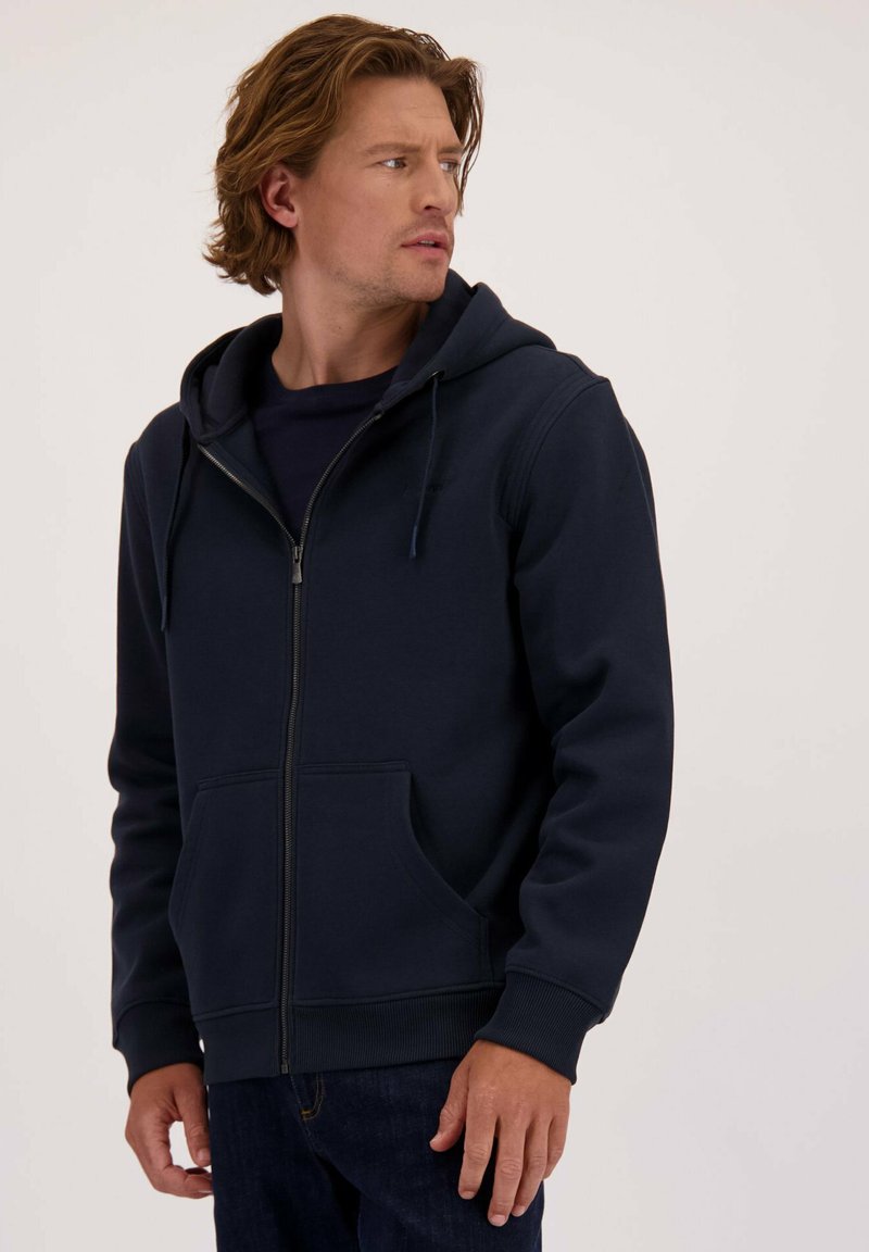 Marineblauwe zip-up hoodie gemaakt van zachte stof, met een kangoeroezak, een capuchon met trekkoord en geribde manchetten. Draagt over een zwart T-shirt.