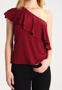 Top burdeos de un solo hombro con un diseño con volantes, hecho de una tela suave, que presenta una silueta ajustada y una textura lisa. Combinado con pantalones negros.