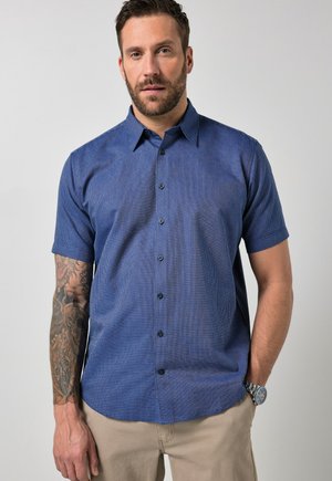 Mann mit Bart trägt ein blaues Kurzarmhemd mit Knöpfen und beige Hosen und zeigt ein detailliertes Tattoo auf seinem linken Unterarm.