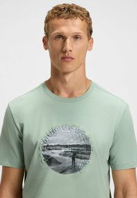 Light green cotton t-shirt featuring a circular graphic with a golfer and "PREMIUM GOLF FEST 1991" text, round neckline, and short sleeves.