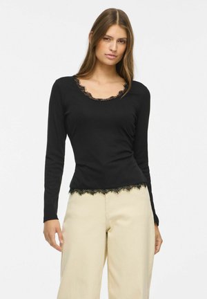 Femme portant un haut à manches longues noir avec bordure en dentelle et un pantalon beige taille haute à jambes larges, debout devant un fond uni.