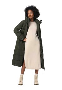 Vadderad lång kappa i mörkgrön med huva, kombinerad med en beige knälång klänning och ljusa snörstövlar.