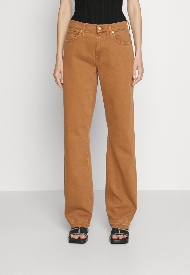 Personne portant un jean marron droit à taille haute, associé à des sandales noires à bout ouvert et un haut noir sans manches, debout devant un fond uni.