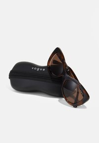 Ochelari de soare tip cat-eye cu model bicolor, alunecând pe o borsetă neagră, cu fermoar, pe care este imprimat brandul "vogue".
