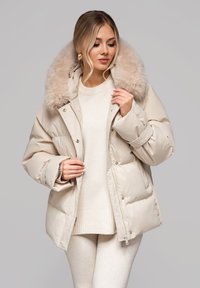Veste matelassée beige avec un grand col en fausse fourrure douce, fermetures à boutons et à glissière, et texture matelassée. Portée sur un pull léger et des leggings.