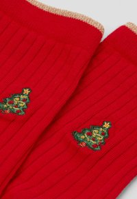 Červené žebrované ponožky se zlatým lemováním, zdobené vyšitými zelenými vánočními stromy s žlutými a červenými detaily.