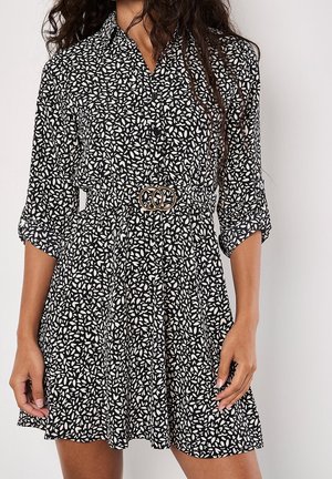 Femme portant une robe noire et blanche à motifs de feuilles, manches retroussées et ceinture à la taille avec deux anneaux dorés entrelacés.