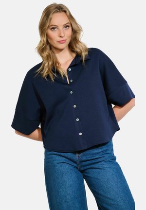 Navy cropped shirt with a button-up front, short sleeves, and a collar. Soft fabric, relaxed fit, and subtle sheen. Paired with blue jeans.