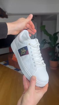 Mains tenant une basket blanche avec des lacets blancs et une bande brodée bleu-rouge en forme de blason sur le côté, en intérieur avec des plantes en arrière-plan.