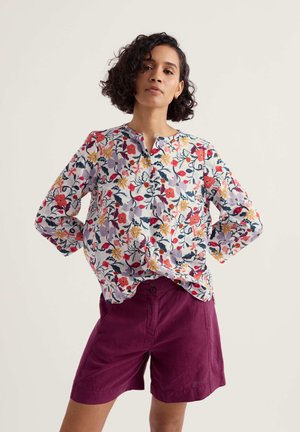 Žena s kudrnatými vlasy, oblečená v květované blůze s dlouhými rukávy a tmavě fialových kraťasech s vysokým pasem, stojící s rukama v bok.