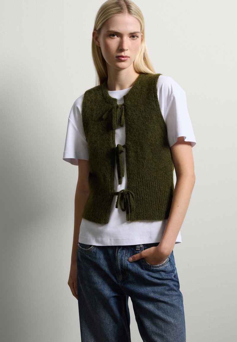 Olijfgroene gebreide vest met voorstriksels, gedragen over een wit T-shirt. Casual outfit met ontspannen blauwe spijkerbroek. Eenvoudig ontwerp.