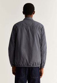 Un adulte portant une veste légère gris foncé avec un ourlet élastique et un pantalon en denim bleu, se tenant de dos face à l'appareil photo.