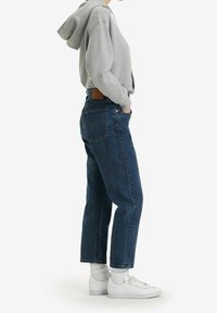 Jean en denim de couleur bleu foncé, coupe droite, avec une étiquette en cuir au dos et des poches arrière. Associé à un sweat à capuche gris et des baskets blanches.