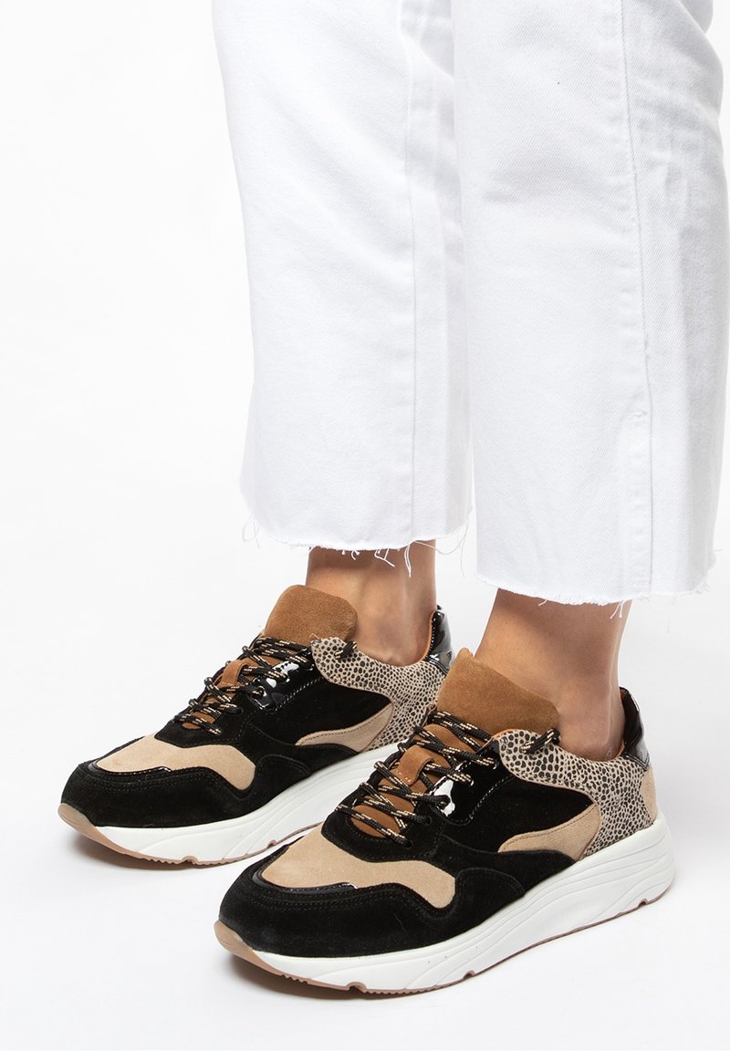 Pieds portant des baskets à motifs noirs, beiges et marron avec des semelles blanches, associées à un pantalon blanc court aux ourlets effilochés.