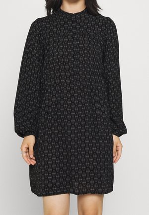 Kobieta w czarnej sukience z długim rękawem o geometrycznym wzorze, stojąca z rękami swobodnie opuszczonymi wzdłuż ciała.
