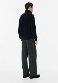 Férfi, aki enyhén elfordulva áll, fekete garbó pulóvert, szürke, bő szárú nadrágot és fekete cipőt visel fehér háttér előtt.