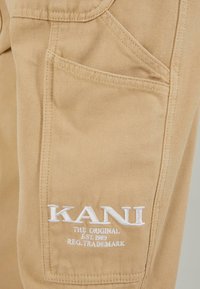 Beige puuvillahousut, joissa on etutasku ja valkoinen brodeerattu "KANI"-logo tekstillä, teksturoitu kangas ja siistit ompeludetalit.